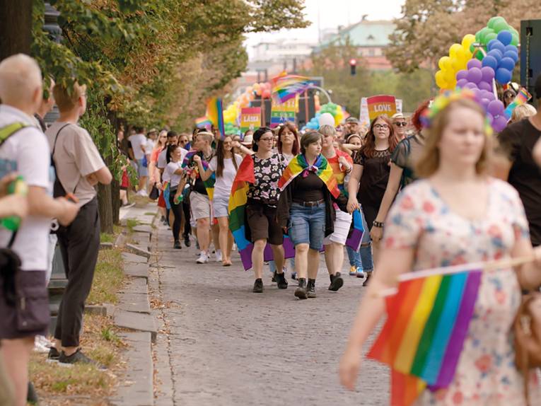 Das Bild zeigt mehrere Menschen bei einer Pride-Parade. Sie tragen Regenbogenfahnen oder halten sie hoch.